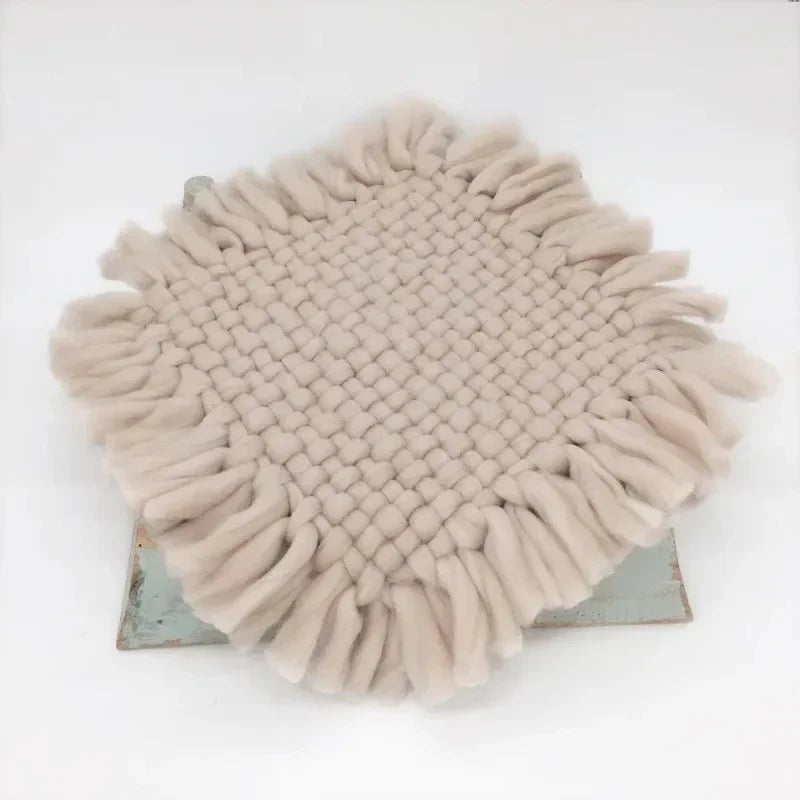 A round woven throw blanket with long fringe trim in a light beige or oatmeal color, displayed on a rustic metal stand against a plain white background.