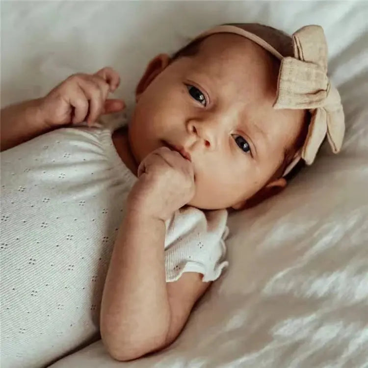 Baby wearing a white outfit and beige headband with a bow, lying on a soft surface.