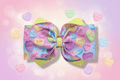 Hero image of pastel candy hearts hair bow on dreamy Valentine's Day inspired background with sparkles