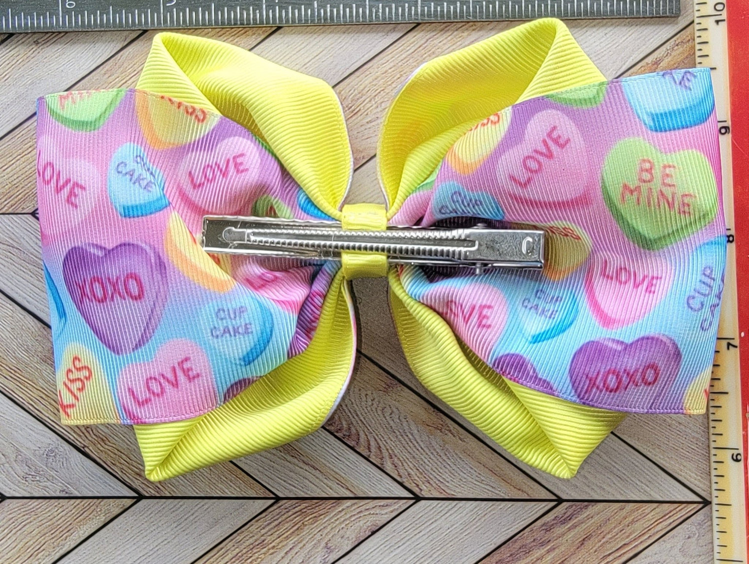 Colorful hair bow with heart patterns on a wooden surface next to a ruler.