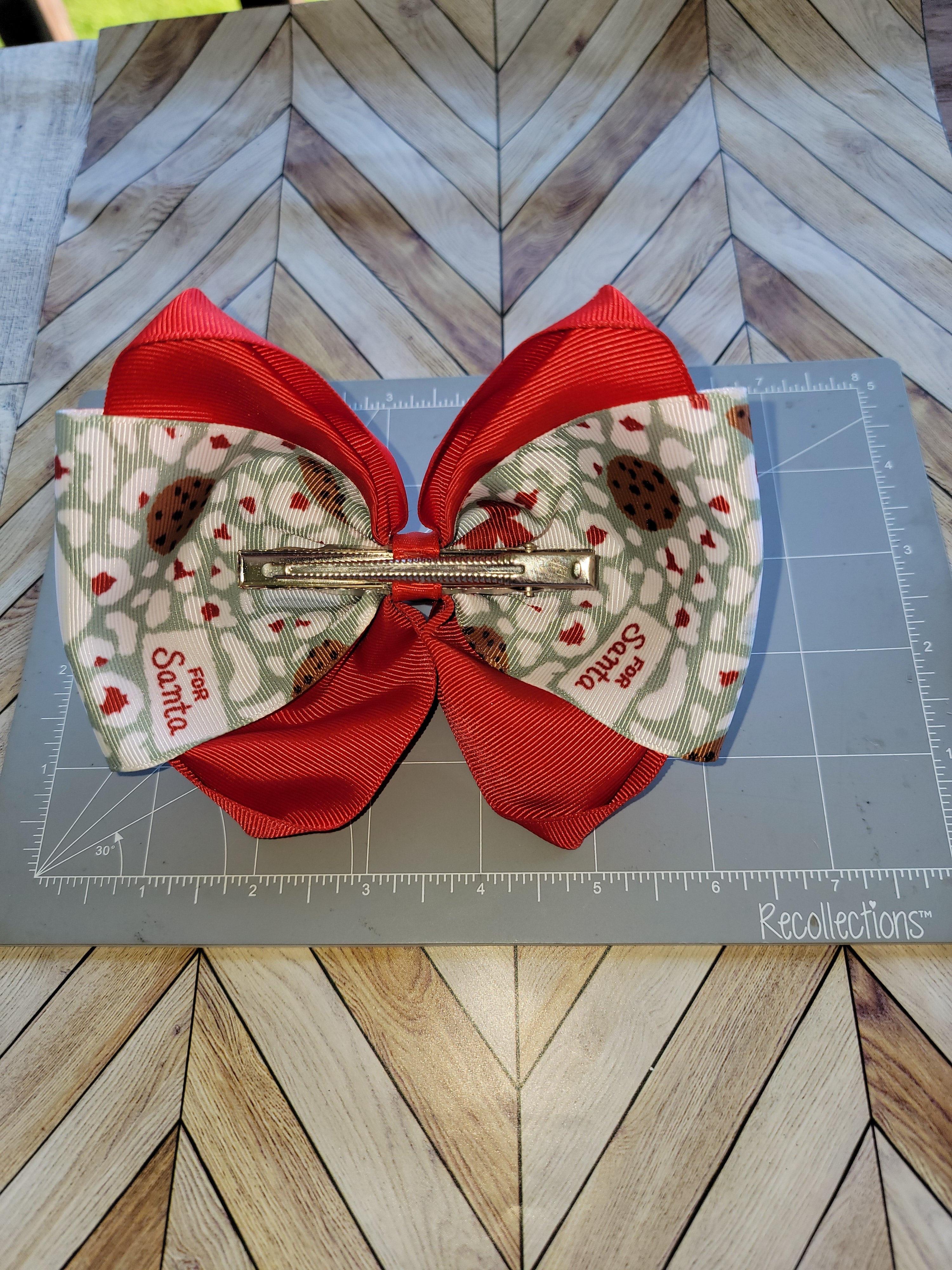 A handmade hair bow accessory featuring red grosgrain ribbon loops and patterned paper wings with a Santa print, secured with a metal claw clip, displayed on a gray cutting mat over a wood chevron background.