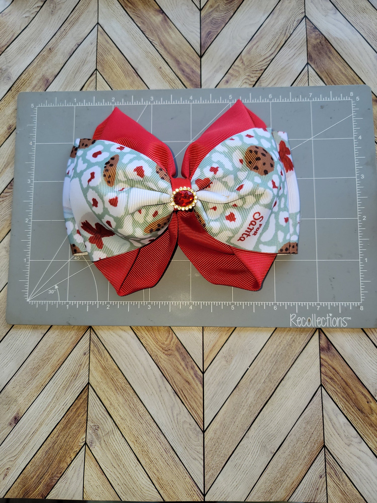A handmade hair bow made from red grosgrain ribbon and patterned fabric featuring Santa and holiday motifs, displayed on a gray cutting mat over a wood herringbone floor.