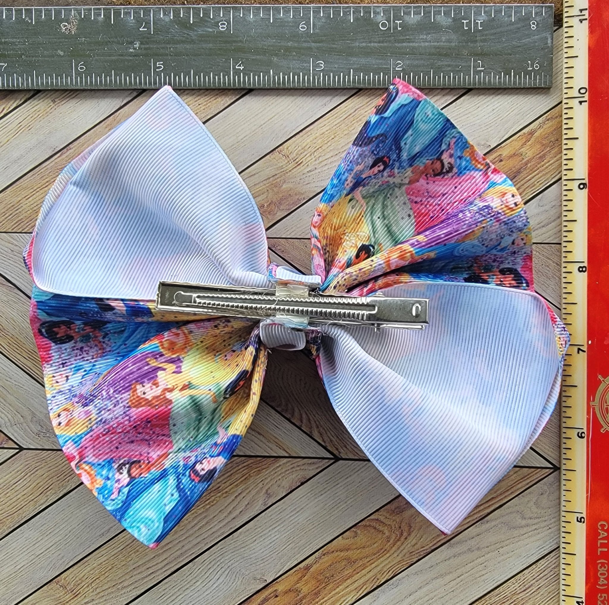 Colorful hair bow with a clip on a wooden surface next to a ruler.