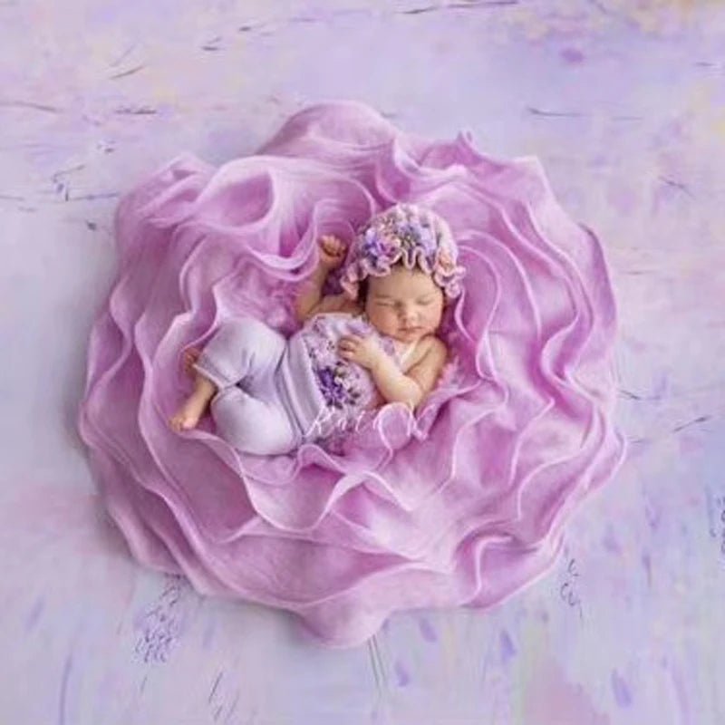 Newborn baby in a floral bonnet and white outfit lying on pink fabric with a soft purple background