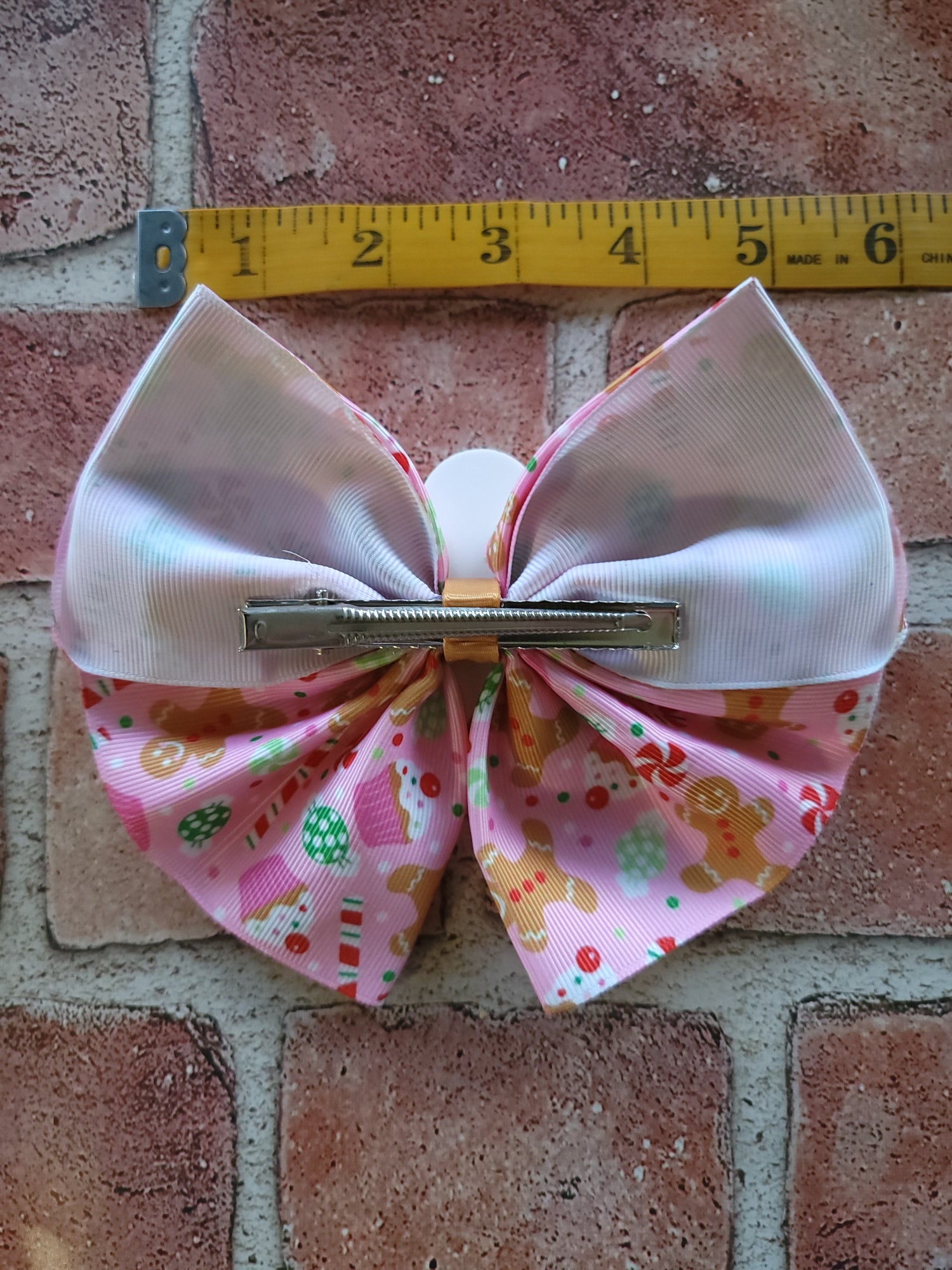 A pink hair bow with a candy and gingerbread print on the bottom half and iridescent white satin on the top half, shown with a yellow measuring tape above it on a brick surface.