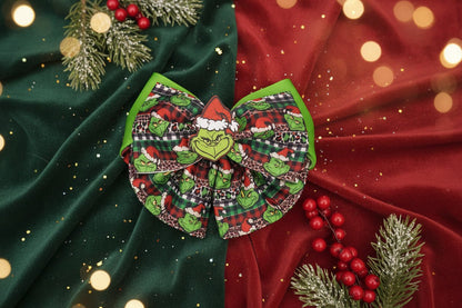A Christmas-themed hair bow featuring a Grinch face, green Grinch characters, red and black plaid patterns, and green trim, displayed on a light gray textured surface.