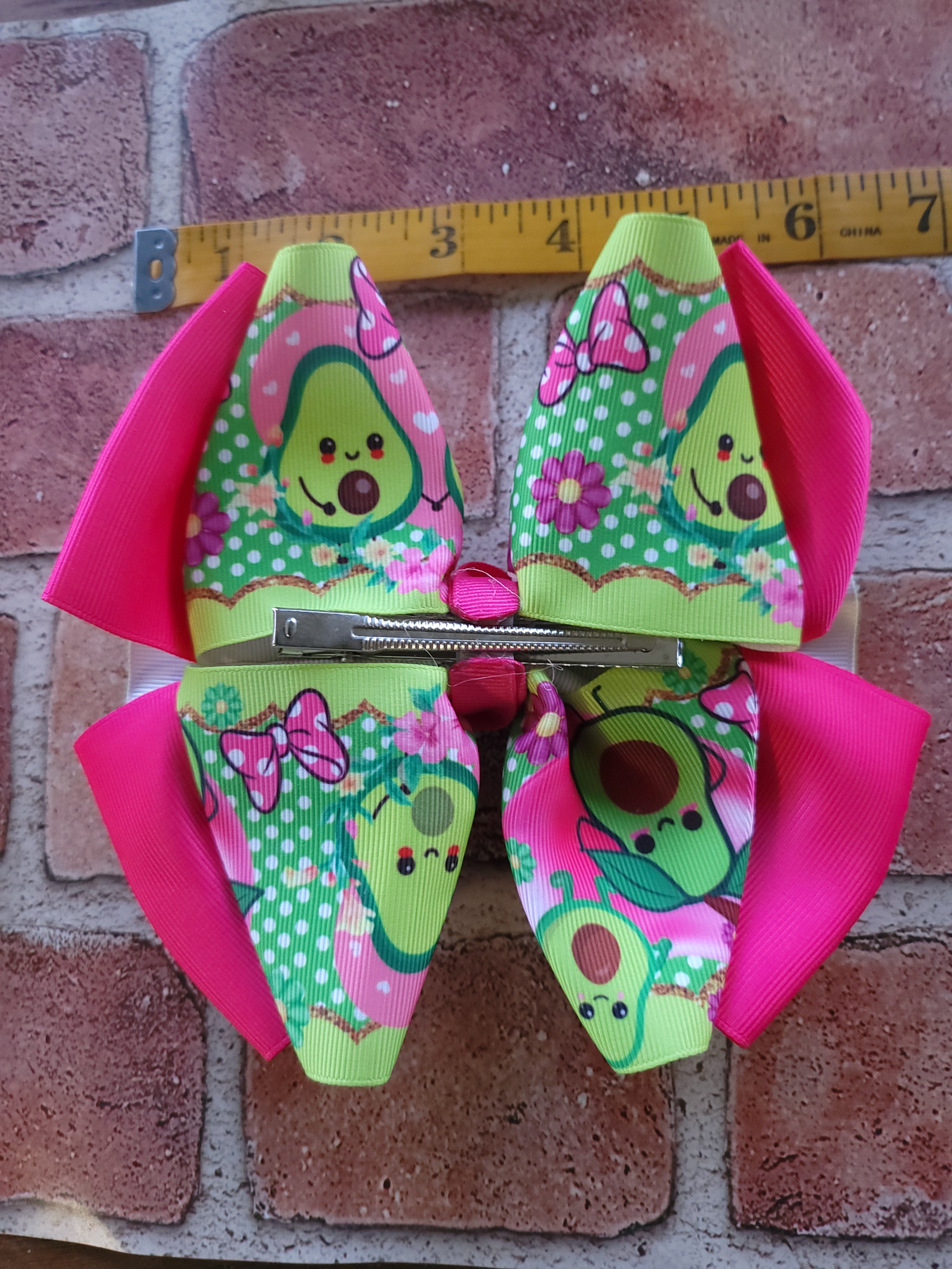 A colorful hair bow with a cartoon avocado pattern, pink trim, and a pink center, displayed on a brick surface next to a yellow measuring tape