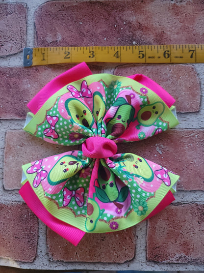 A colorful hair bow with a cartoon avocado pattern, pink trim, and a pink center, displayed on a brick surface next to a yellow measuring tape.