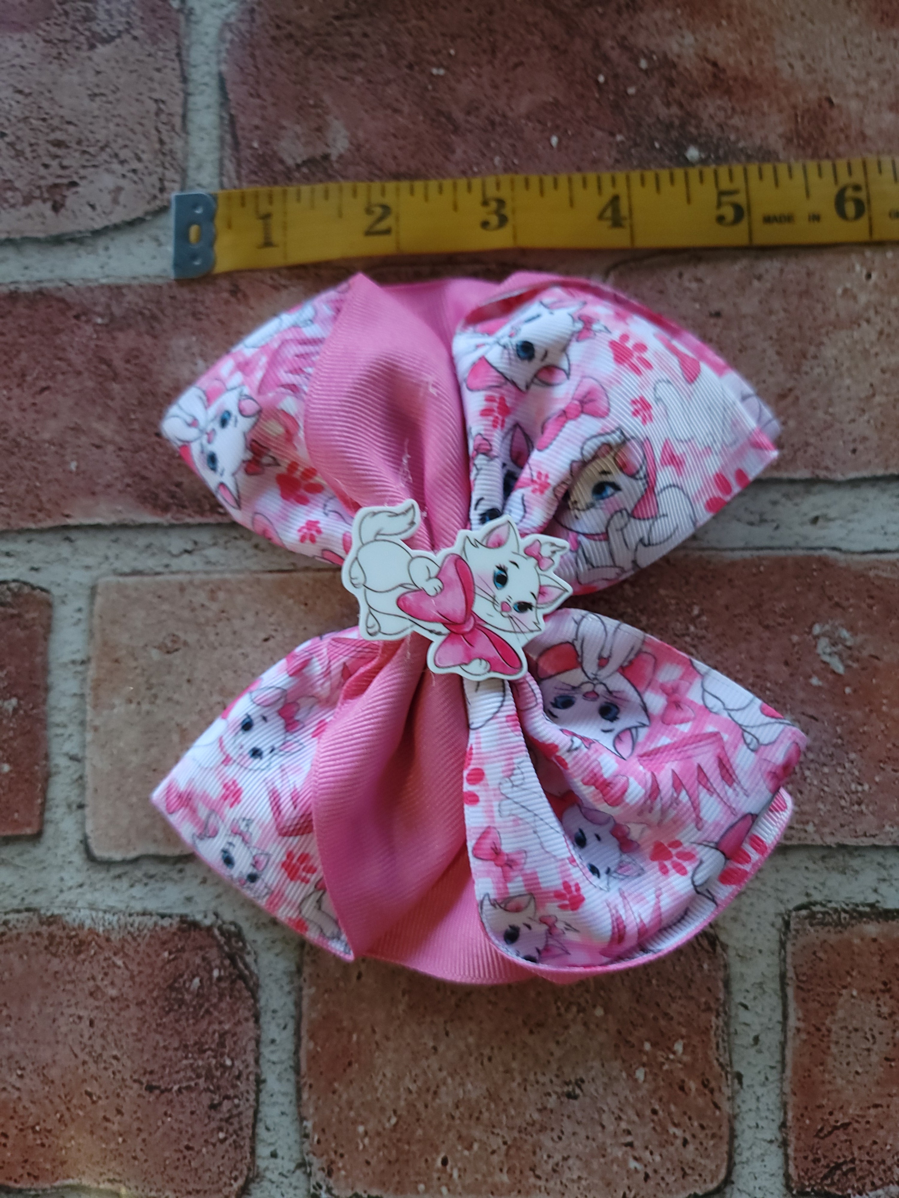 A pink hair bow with a white and pink cartoon cat pattern, a solid pink ribbon center, and a Marie Antoinette charm, shown next to a yellow measuring tape on a brick background.