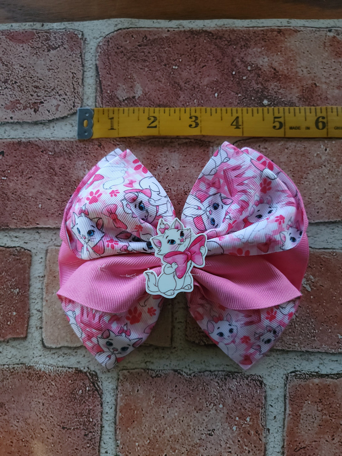 A pink and white patterned hair bow with a cartoon character in the center, placed on a brick surface next to a yellow measuring tape.