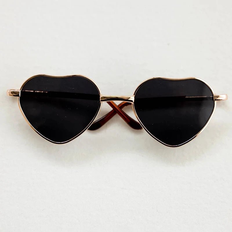 Heart-shaped sunglasses with black lenses and gold frames on a light gray background