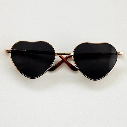 Heart-shaped sunglasses with black lenses and gold frames on a light gray background