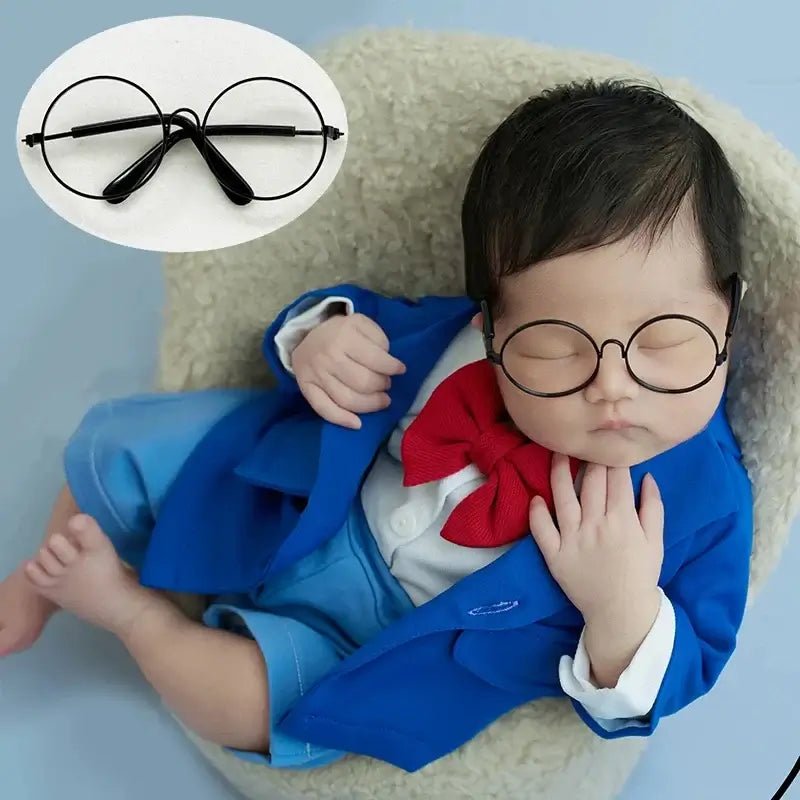 Baby wearing a blue outfit with a red bow tie and black glasses, lying on a light-colored surface.