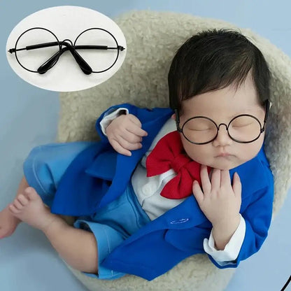 Baby wearing a blue outfit with a red bow tie and black glasses, lying on a light-colored surface.