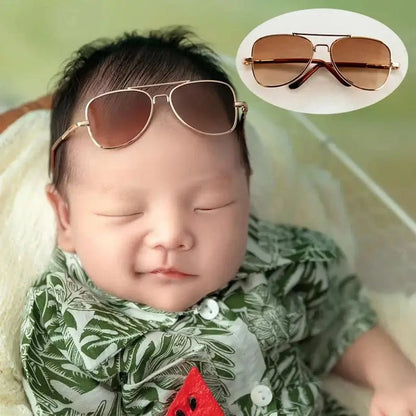 Baby wearing sunglasses with a close-up inset of sunglasses, set against a blurred green background.