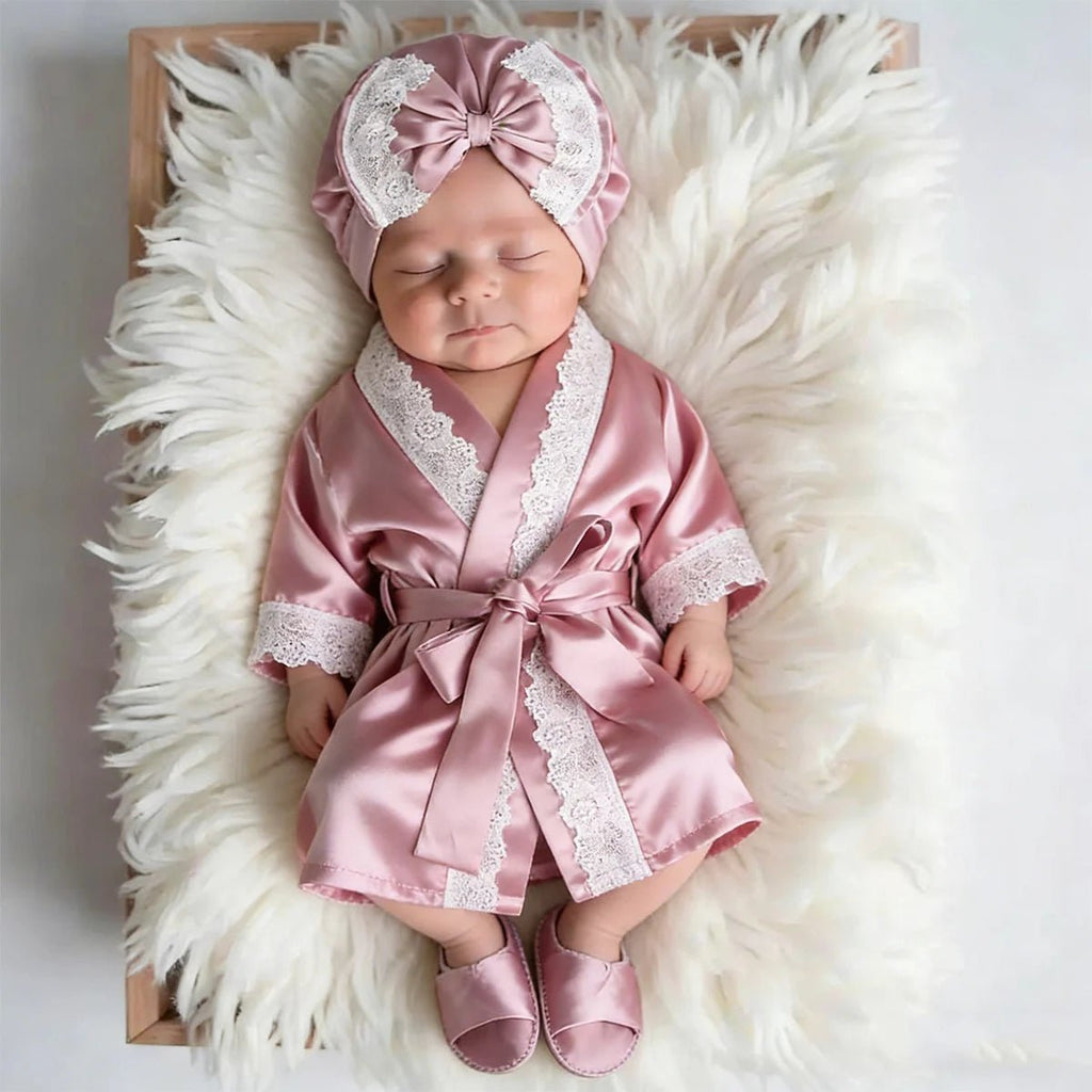 Newborn baby in a pink satin robe and matching headband on a fluffy white surface.