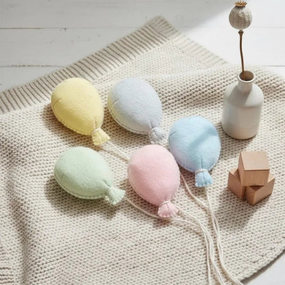 Pastel-colored plush balloons on a textured surface with a vase and wooden block in the background.