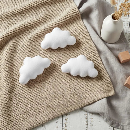 White cloud-shaped cushions on a textured fabric surface with a vase and wooden blocks in the background.