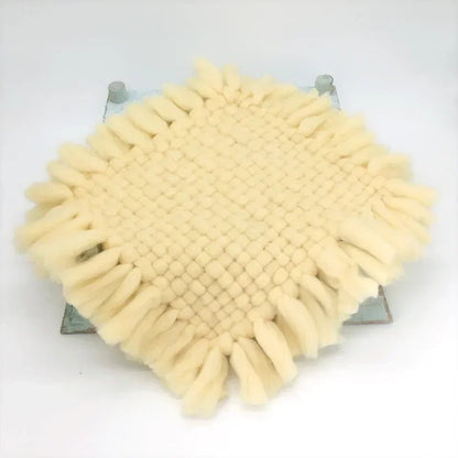 A square woven textile pad in a warm cream or pale yellow color, featuring a textured grid pattern and long fringe along all four edges, displayed on a light gray background.