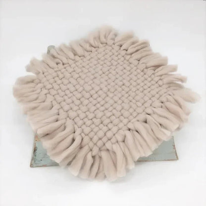A round woven throw blanket with long fringe trim in a light beige or oatmeal color, displayed on a rustic metal stand against a plain white background.