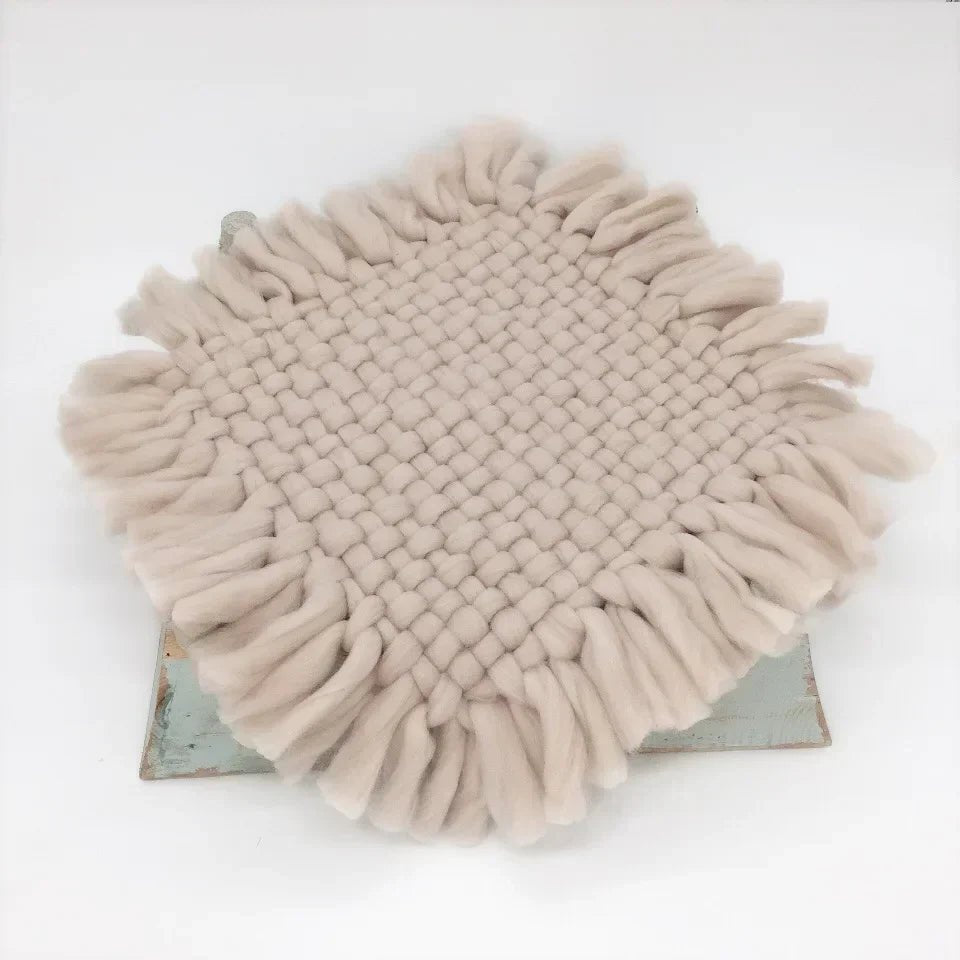 A round woven throw blanket with long fringe trim in a light beige or oatmeal color, displayed on a rustic metal stand against a plain white background.