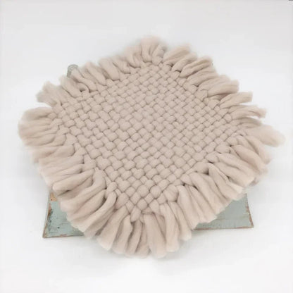 A round woven throw blanket with long fringe trim in a light beige or oatmeal color, displayed on a rustic metal stand against a plain white background.