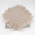A round woven throw blanket with long fringe trim in a light beige or oatmeal color, displayed on a rustic metal stand against a plain white background.