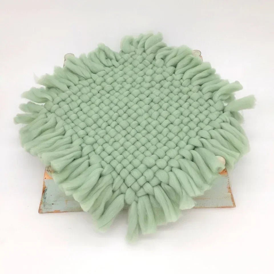 A round sage green woven fringed doily or coaster displayed on a small rustic wooden board against a plain white background.