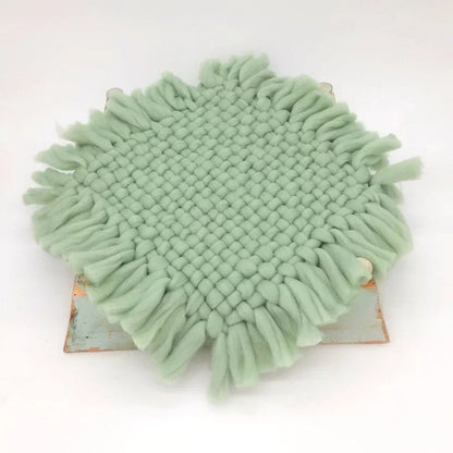 A round sage green woven fringed doily or coaster displayed on a small rustic wooden board against a plain white background.