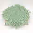 A round sage green woven fringed doily or coaster displayed on a small rustic wooden board against a plain white background.