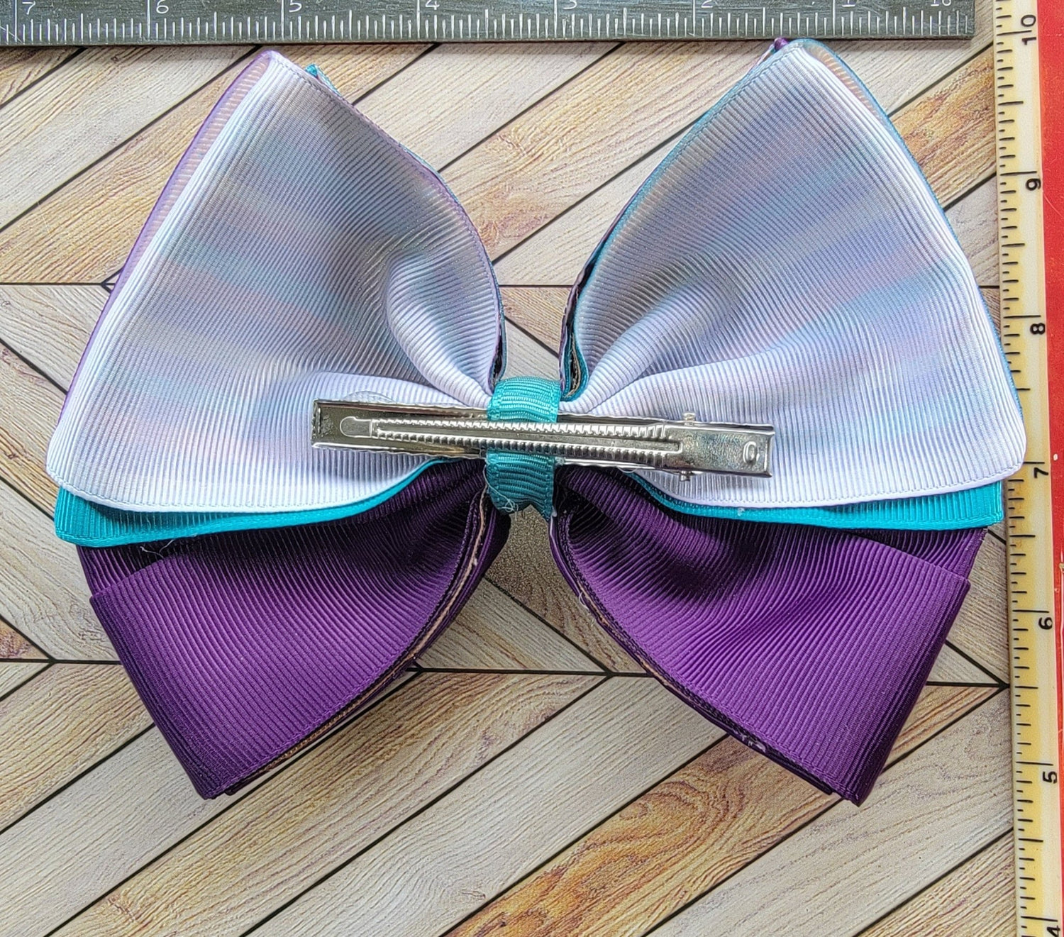 Large hair bow with purple and white sections on a wooden surface with a ruler for scale.
