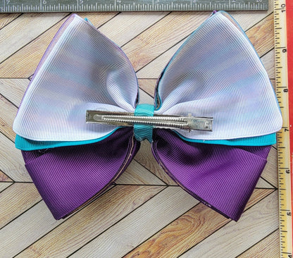 Large hair bow with purple and white sections on a wooden surface with a ruler for scale.