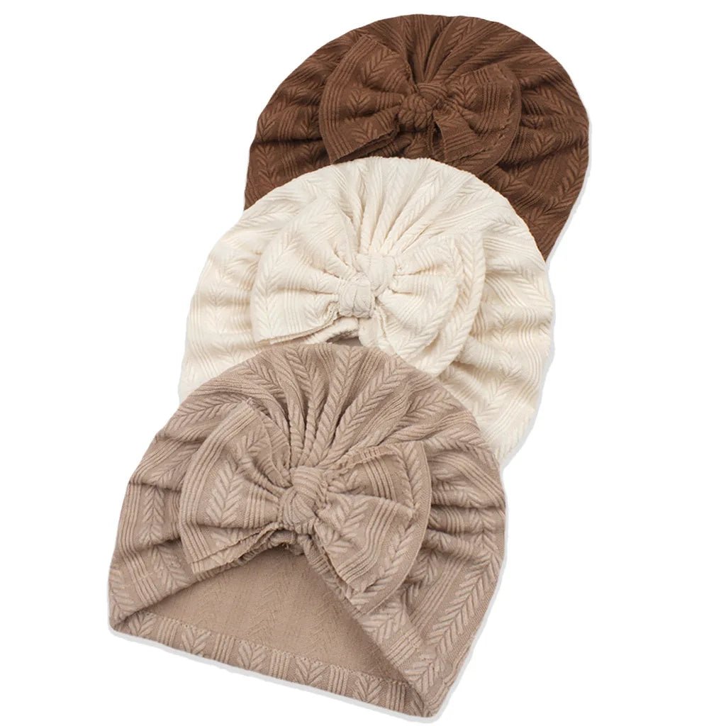Three baby turbans in brown, beige, and white with bow details on a white background.