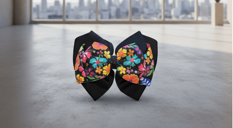 A handmade hair bow made of black grosgrain ribbon and fabric panels with a vibrant multicolor floral print, displayed on a gray cutting mat over a wood herringbone floor.