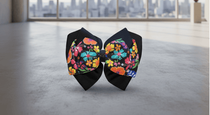 A handmade hair bow made of black grosgrain ribbon and fabric panels with a vibrant multicolor floral print, displayed on a gray cutting mat over a wood herringbone floor.