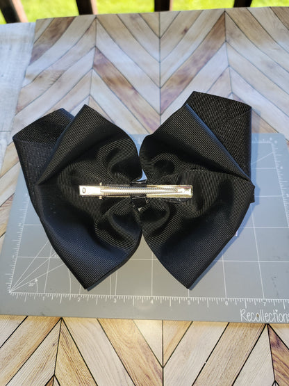 A large black double-bow hair clip with a silver metal barrette clasp, laid flat on a gray cutting mat over a wood-patterned surface.