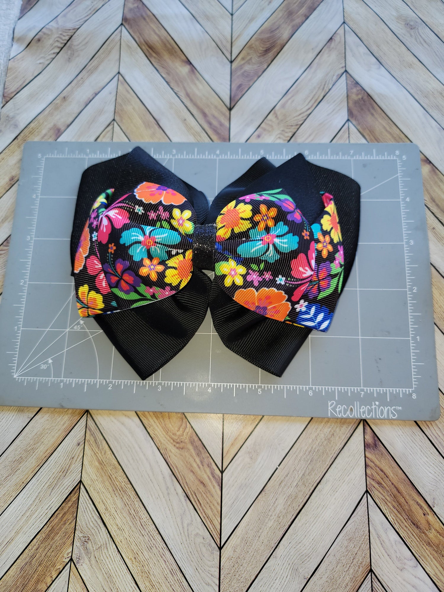 A handmade hair bow made of black grosgrain ribbon and fabric panels with a vibrant multicolor floral print, displayed on a gray cutting mat over a wood herringbone floor.
