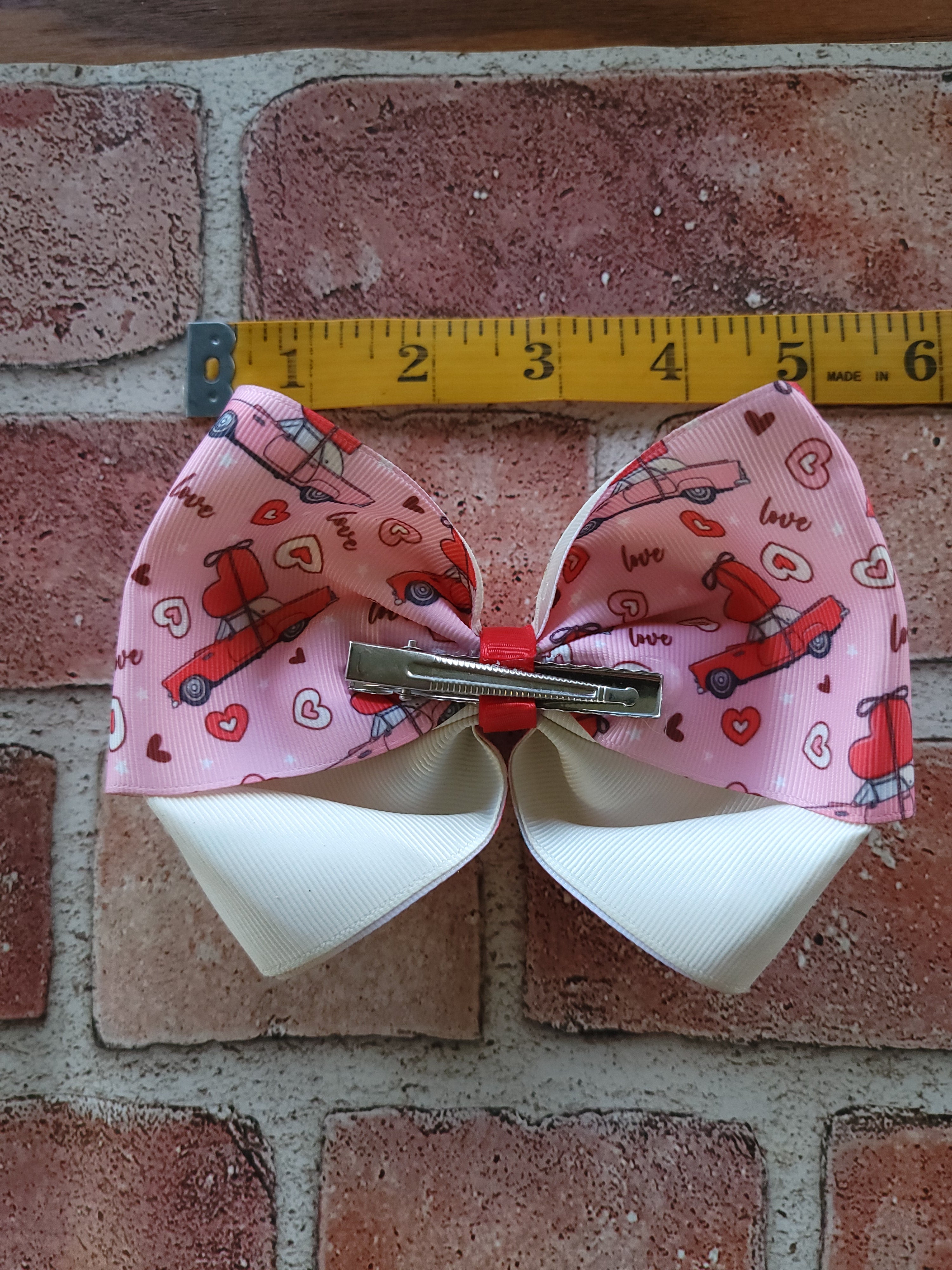 A pink hair bow with a red double-decker bus pattern and white reverse sides, shown next to a yellow measuring tape on a brick surface.