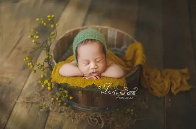 Newborn baby sleeping in a wooden bucket with green bonnet and yellow blanket, L&