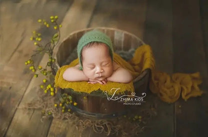 Newborn baby sleeping in a wooden bucket with green bonnet and yellow blanket, L&