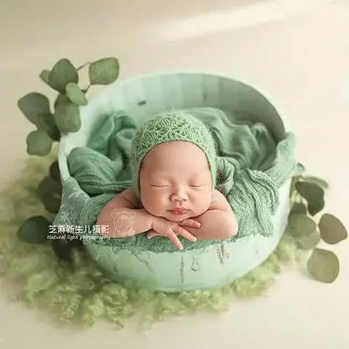 Newborn baby in a green outfit lying in a green bowl with greenery around.
