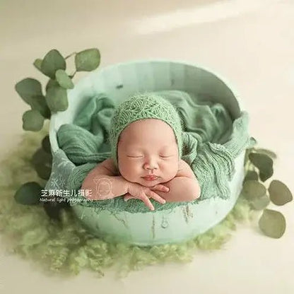 Newborn baby in a green outfit lying in a green bowl with greenery around.