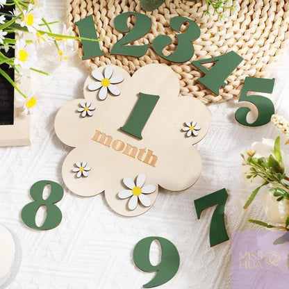 Wooden month calendar numbers arranged on a white textured fabric with daisy flowers and a woven placemat. The central flower-shaped plaque reads &
