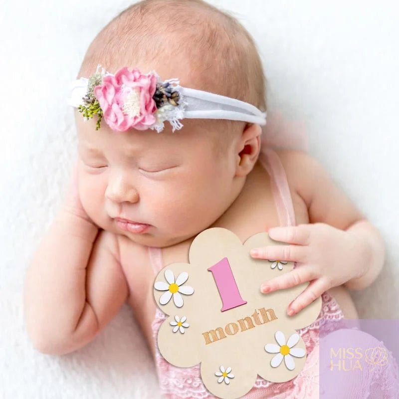A sleeping newborn baby wearing a white fabric headband decorated with a pink flower and small green leaves, holding a decorative &