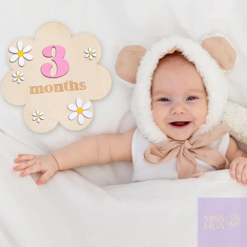 A smiling baby lying on a white blanket wearing a white and beige bunny-ear bonnet with a large beige bow. A decorative cloud-shaped sign reads &