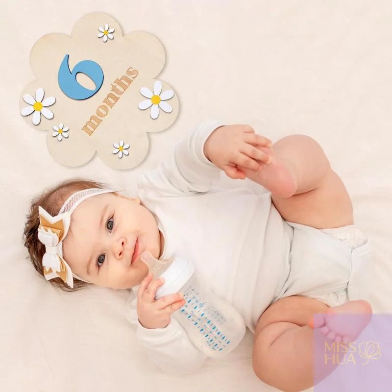 A smiling baby lying on a white blanket wearing a white large beige bow. A decorative cloud-shaped sign reads &