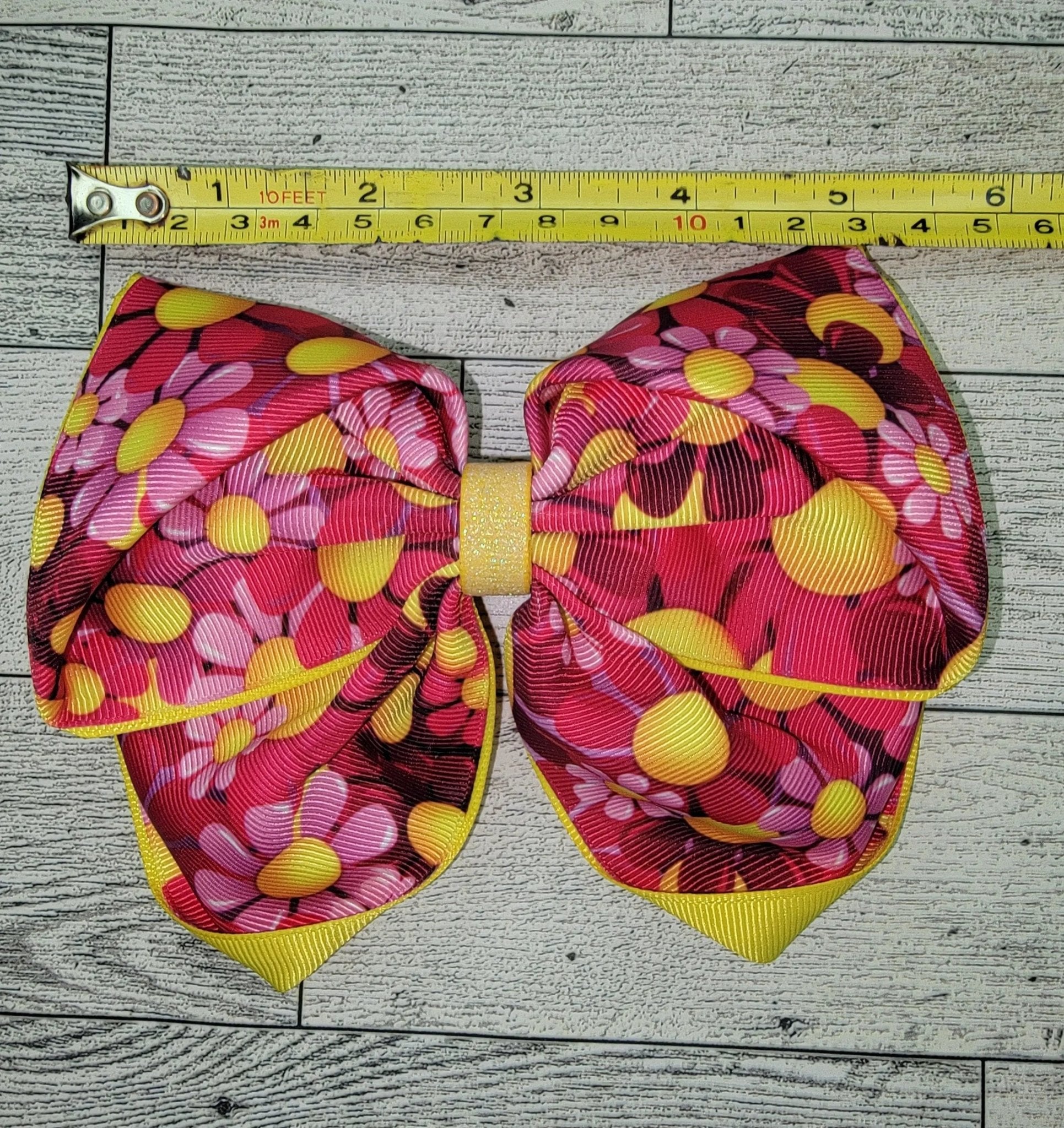 Large decorative bow with pink and yellow pattern on a wooden surface, measured by a tape measure.